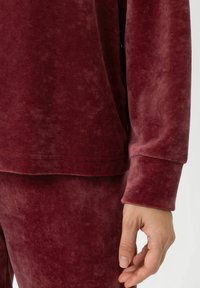 Bordeauxvelours top met lange mouwen, met een gladde textuur en brede manchetten; gedragen met bijpassende onderstukken, wat een gecoördineerde outfit laat zien.