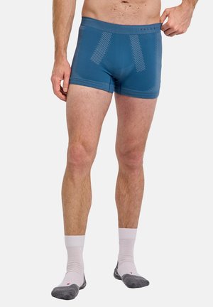 Man draagt blauwe boxer en witte sokken met grijze tenen, staat tegen een effen achtergrond en houdt de tailleband aan één zijde vast.