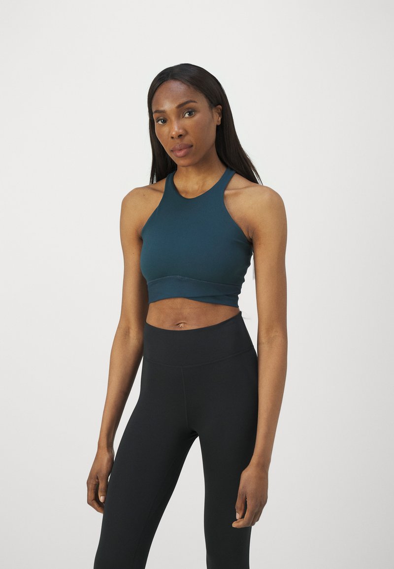 Top desportivo cropped em verde escuro com um design de halter neck, emparelhado com leggings pretas de cintura alta. Textura suave e detalhes mínimos.