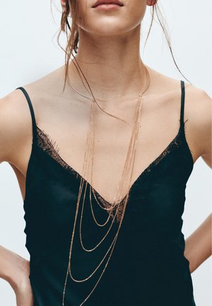 Nő fekete csipkés szegélyű camisole-t visel, mellkasán hosszú, réteges aranylánc nyaklánccal.