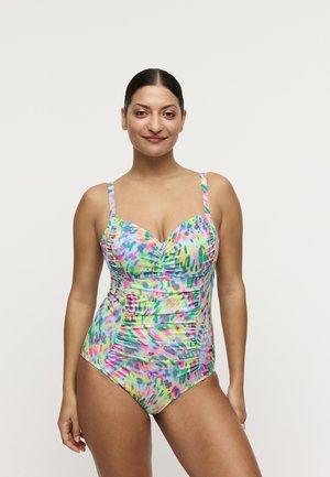 Vrouw staand en glimlachend, gekleed in een kleurrijk patroon badpak met geplooide details, tegen een effen lichte achtergrond.