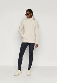 Bēšs flīsa kapuce ar priekšējo kabatu, kopā ar tumšām legingām un baltām sporta kurpēm. Modelis valkā saulesbrilles un viņai ir pītas matu frizūra.