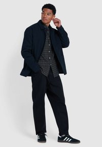Veste bleu foncé sur une chemise à carreaux à boutons, pantalon noir et baskets noires. Les matériaux semblent robustes avec une coupe décontractée et des lignes épurées.