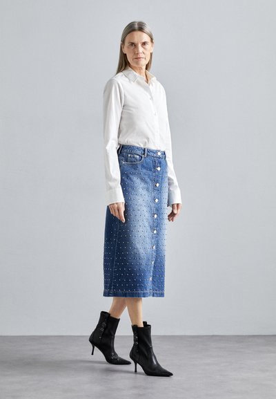 Jeans-Midirock mit silberner Knopfverschluss, genieteter Textur und leichtem Verblassen. Kombiniert mit schwarzen Knöchelstiefeln und einem weißen Hemd mit Knopfleiste.