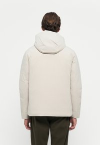 Giacca puffer beige chiaro con cappuccio, caratterizzata da una superficie liscia e polsini elasticizzati. Il design include una vestibilità dritta e uno stile minimalista.