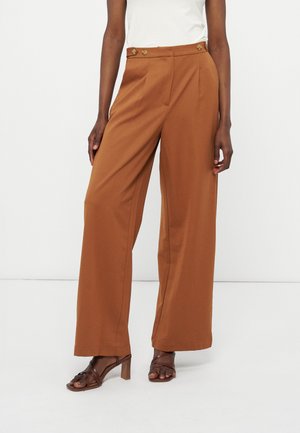 Γυναίκα που φοράει ψηλόμεσα, φαρδιά παντελόνια σε σκουριά απόχρωση με λεπτομέρειες κουμπιών και καφέ πέδιλα με τακούνια.