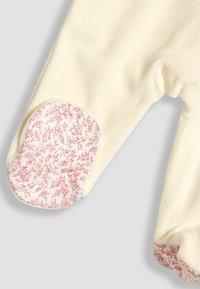 Cremfarbener Overall aus glattem Stoff; rosa Blumenakzente an Ärmeln und Füßen für zusätzliche Details. Verfügt über abgerundete Fußpatches.