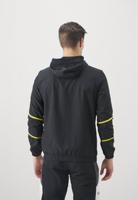 Chaqueta ligera negra con capucha, con detalles amarillos en las mangas y una cinturilla elástica. Textura suave y diseño casual.