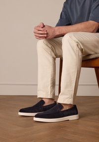 Homme portant un pantalon beige et des chaussures noires sans lacets assis sur une chaise en bois, les mains croisées sur le genou, sur un sol en bois.
