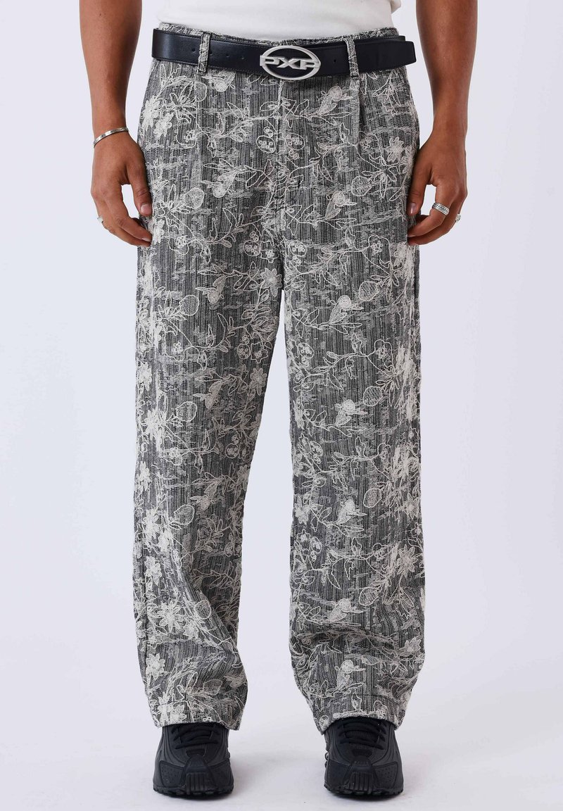 Homme portant un pantalon gris texturé avec une broderie florale blanche, une ceinture noire avec une boucle ovale, des baskets noires, ainsi que des bagues et un bracelet argentés.