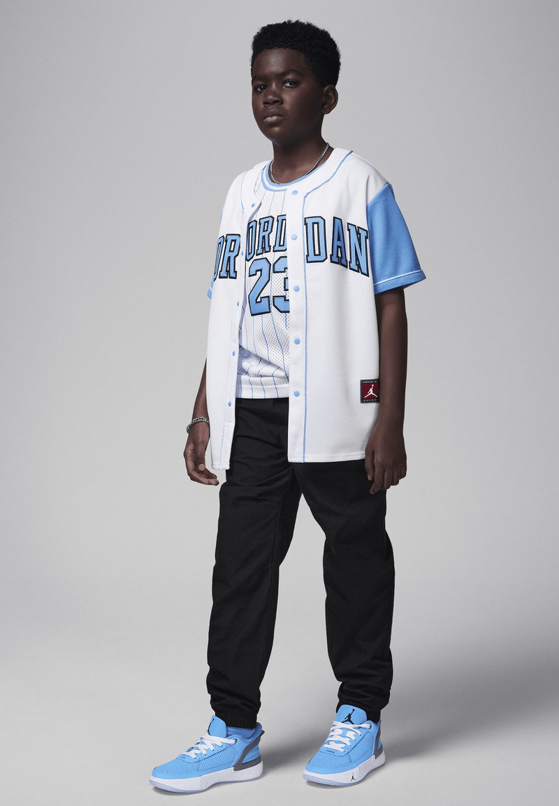 A fiatal egy fehér mezben van, kék díszítéssel és "Jordan 23" felirattal, fekete nadrággal és világoskék sportcipővel párosítva. Texturált, sportos dizájn.
