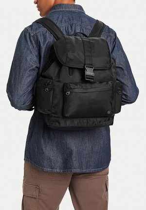 Uomo visto da dietro che indossa uno zaino nero con patta frontale e molte tasche con cerniera, camicia di denim e pantaloni cargo marroni.