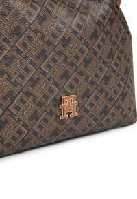 Σκούρο καφέ τσάντα με σχέδιο και χρυσαφί διακοσμητικό λογότυπο "TH" στο κάτω κέντρο, σε λευκό φόντο.