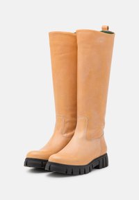 Botas de cano alto em couro castanho com textura suave, bico redondo e sola de borracha robusta. Apresentam um laço para facilitar o calçar.