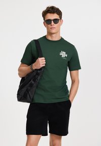 Zelené bavlnené tričko s bielym textom, čierne kordové šortky a čierna taška. Okruhé slnečné okuliare dotvárajú celý outfit.