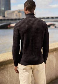 Tricou negru tricotat cu un model texturat, guler înalt și manșete riguroase, asortat cu pantaloni de culoare deschisă, cu o suprafață netedă.
