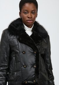 Veste en cuir noir avec un col en fausse fourrure, design à double boutonnage, boutons rehaussés d'or et texture matelassée sur les manches.