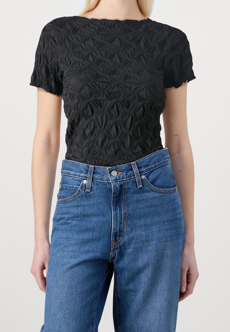 Femme portant un haut noir texturé à manches courtes et un jean bleu taille haute, debout devant un fond uni et clair.