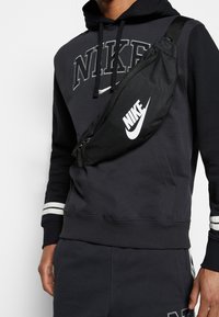 Svart midjeväska från Nike med vitt logo, tillverkad av tåligt tyg. Har en böjd form, fack med dragkedja och justerbar rem.