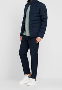 Marineblå puffjacka med knappdetalj, två framfickor, bärs över en ljusgrön t-shirt och mörka byxor, matchas med vita sneakers.