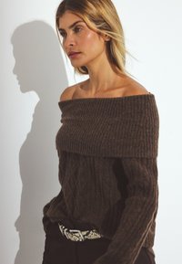 Ruskea olkaimeton villapaita, jossa on ribbinen kaulus ja teksturoitu kuvio. Yhdistetty tummiin housuihin ja kuvioituun vyöhön. Pehmeä, paksu neulottu.