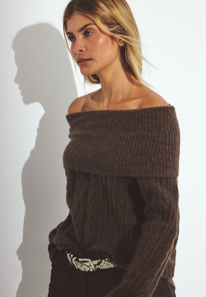 Bruine off-the-shoulder trui met een ribbelkraag en een gestructureerd patroon. Gecombineerd met donkere broeken en een patroongriem. Zachte, chunky brei.