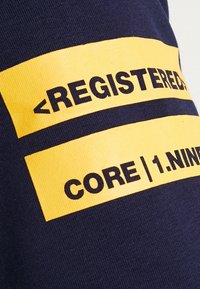 Tela azul marino con dos franjas horizontales amarillas y texto en negro: "<REGISTRADO>" y "CORE | 1.NUEVE." El texto es negrita y prominente.