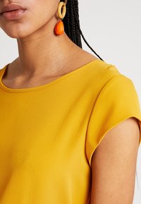 Donna che indossa un top testurizzato giallo senape e un orecchino a goccia arancione con dettagli in legno.