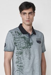 Siva polo majica z darko ovratnikom, zelenim grafičnim besedilom, teksturirano blago in majhnim žepkom na prsih z detajli, podobnimi denimu.