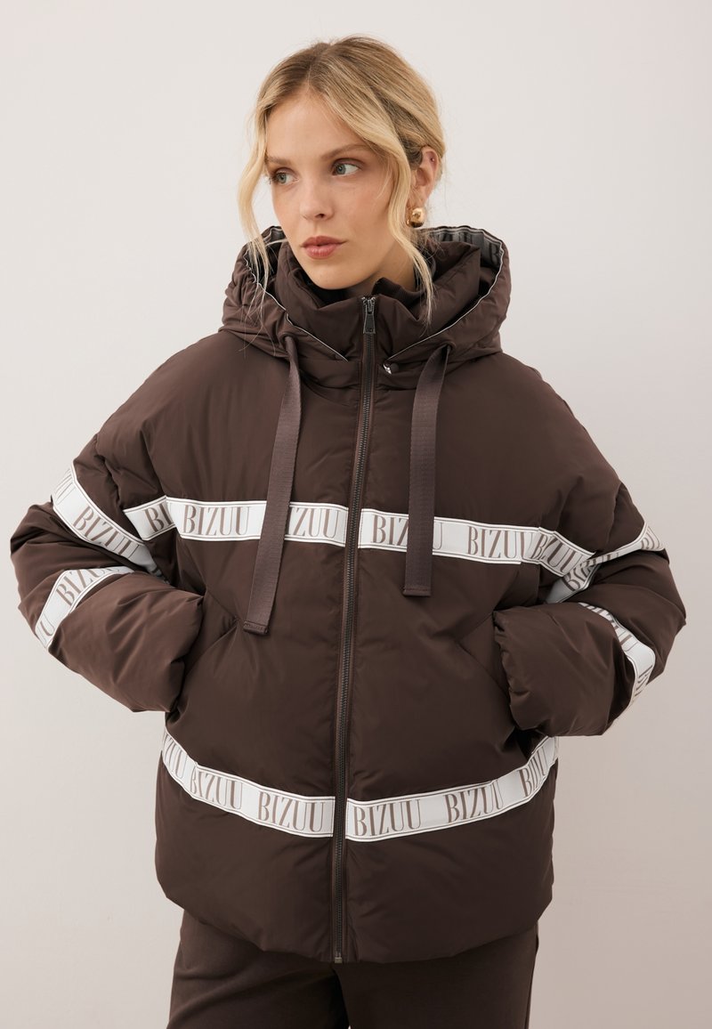 Kobieta ubrana w ciemno brązową kurtkę puchową z kapturem i białymi poziomymi paskami z napisem "BIZUU," z rękami w kieszeniach, patrzy w bok.