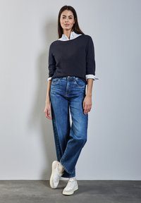 Maglione navy indossato sopra una camicia bianca con colletto, abbinato a jeans larghi blu e sneakers bianche. Texture morbida, vestibilità casual.