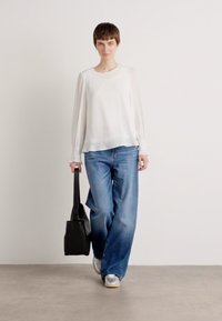 Witte blouse met lange mouwen en doorzichtige mouwen, gecombineerd met lichtblauwe wijde jeans en een zwarte totebag. Model draagt zilveren sneakers.