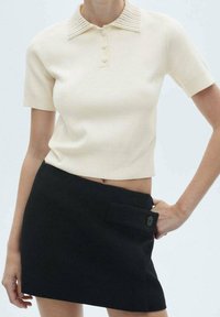 Frau in einem kurzärmeligen, cremefarbenen Strickpolohemd und einem schwarzen asymmetrischen Minirock mit einem geknöpften Klappdetail.