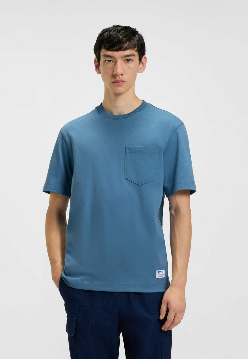 T-shirt blu a maniche corte realizzato in tessuto morbido, con taschino frontale e scollatura rotonda. Indossato con pantaloni scuri.