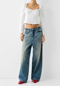 Blusa bianca corta con maniche a sbuffo e bordino in pizzo abbinata a jeans a gamba larga sbiaditi di colore blu e scarpe rosse con tacco alto.