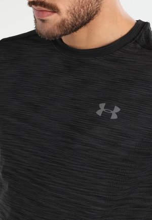 Nahaufnahme des unteren Gesichts und Halses eines Mannes, der ein schwarzes, strukturiertes Under Armour Shirt trägt, mit sichtbarem grauem Logo auf der Brust.
