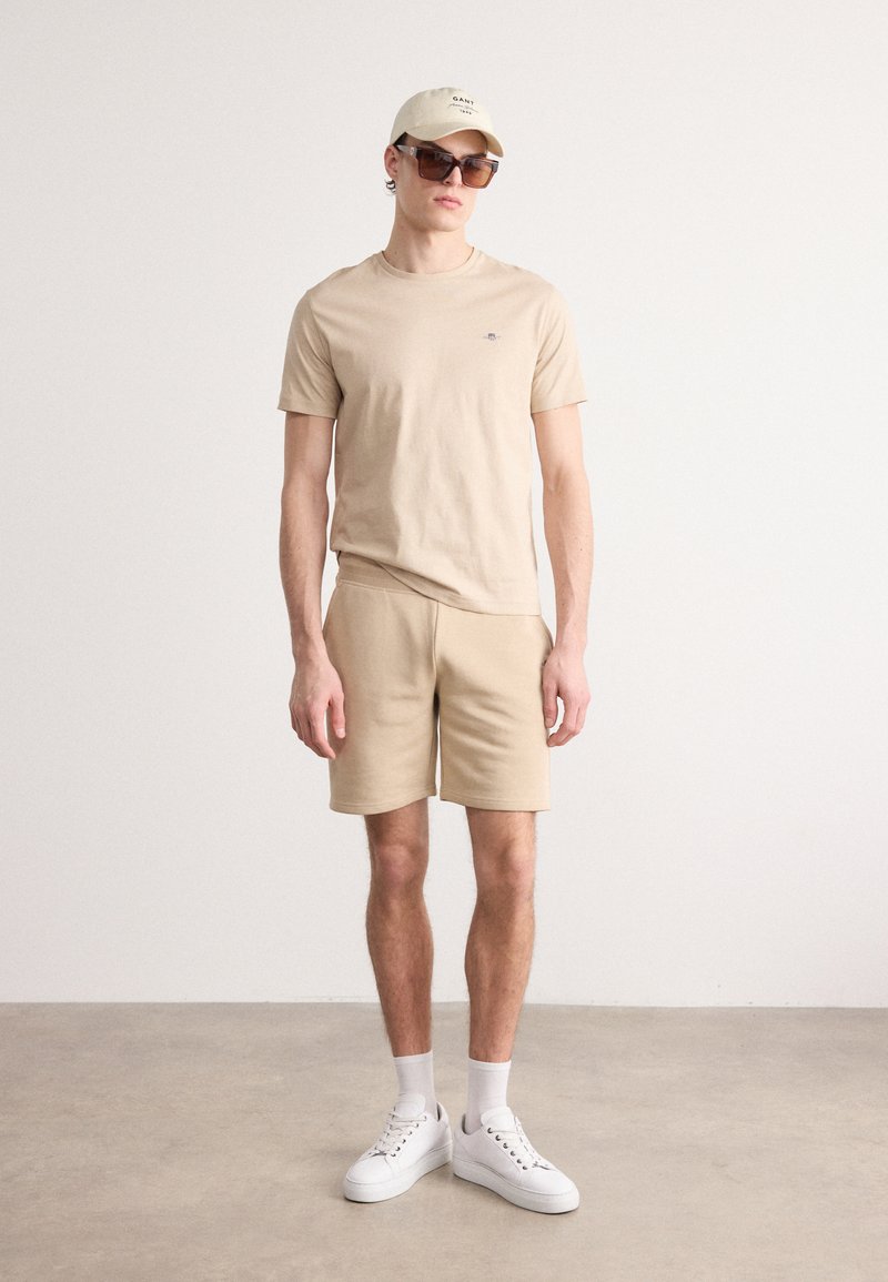 Beige Baumwoll-T-Shirt und Shorts mit einem minimalistischen Design. Die Accessoires umfassen eine leichte Mütze und eine braune Sonnenbrille, kombiniert mit weißen Sneakers.
