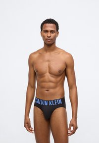 Μαύρα μποξεράκια Calvin Klein με φαρδιά μπλε λογότυπη ζώνη. Το ύφασμα φαίνεται λείο και ελαστικό, σχεδιασμένο για στενή εφαρμογή.