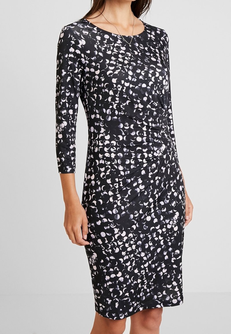 Femme portant une robe noire longueur genou avec un motif abstrait de points blancs et des manches trois-quarts sur fond uni.