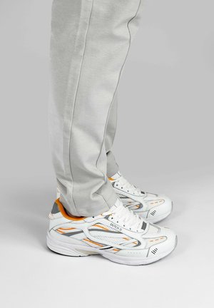 Persoon die lichtgrijze broek en witte hardloopschoenen met oranje en grijze accenten draagt, staand op een effen lichte achtergrond.