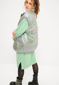 Iridescente gewatteerde vest met hoge kraag, gedragen over een lichtgroene sweaterjurk. Zwarte enkellaarsjes met kettingdetail.