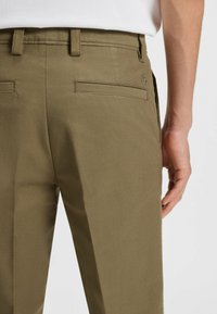 Pantalons vert olive en tissu texturé, avec deux poches arrière et un détail de logo. Design slim-fit avec passants de ceinture.