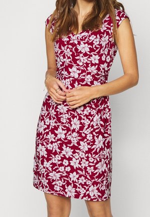 Frau trägt ein knielanges, kurzärmeliges rotes Kleid mit weißem Blumenmuster und steht mit gefalteten Händen vor einem schlichten Hintergrund.