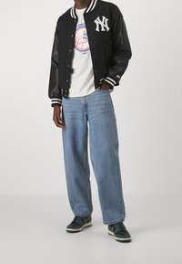 Man i svart och vit varsityjacka med "NY"-logotyp, vit Yankees-t-shirt, vida blå jeans och svarta och gröna sneakers, stående.