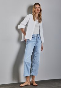 Biała koszula zapinana na guziki noszona na gładkim białym topie na ramiączkach, w połączeniu z jasnoniebieskimi, szerokimi dżinsami. Gładki materiał, luźny krój i skrócona długość.