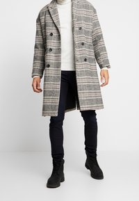 Abrigo a cuadros en gris con acentos en negro y rojo, diseño de doble botonadura y grandes botones. Lleva puesto sobre un suéter ligero y jeans oscuros.