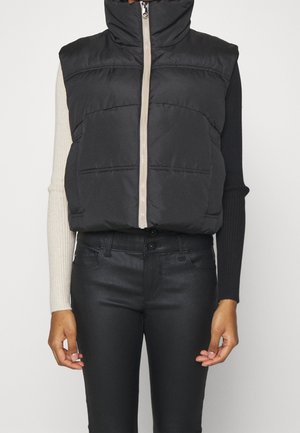 Person trägt eine schwarze ärmellose gesteppte Weste mit Frontreißverschluss über einem langärmligen Oberteil mit kontrastierenden schwarzen und beigen Ärmeln.