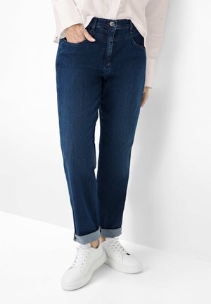 Person iført mørkeblå opsmøgede jeans, lyserød langærmet skjorte og hvide sneakers, stående mod en hvid baggrund.