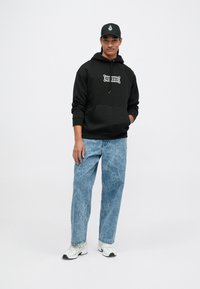 Młody mężczyzna w czapce z daszkiem, czarnej bluzie z kapturem z białym logo, jasnoniebieskich dżinsach typu acid-wash i białych sneakersach na jednolitym białym tle.