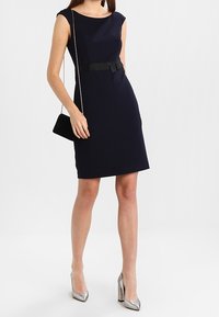 Marinblå klänning med kjol som har en figursydd silhuett, korta ärmar och ett svart bälte som accent. Matchas med silverfärgade högklackade skor och en liten svart väska.